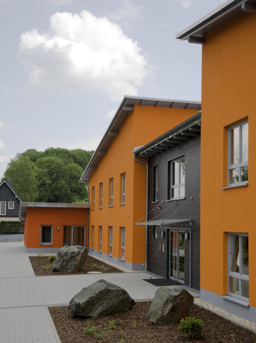 Die eingeschossigen Eckgebäude mit dem dunklen orangeroten Farbton wirken wie eine Einfassung und lassen die einzelnen Baukörper als ein geschlossenes Ganzes erscheinen.