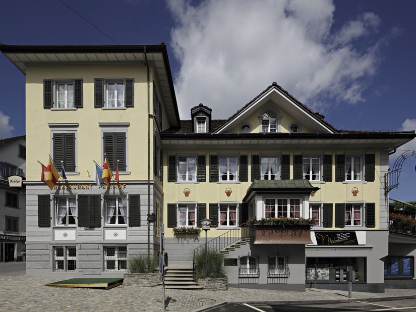 Die drei eingesetzten Farbtöne dominieren die Fassade. Im Sockelbereich wurde mit zwei Grautönen gearbeitet, bei den oberen Fassadenflächen mit einem Gelbton und die Fensterläden bilden mit ihrem dunkeln Farbton den Übergang zum ebenfalls dunklen Dach.