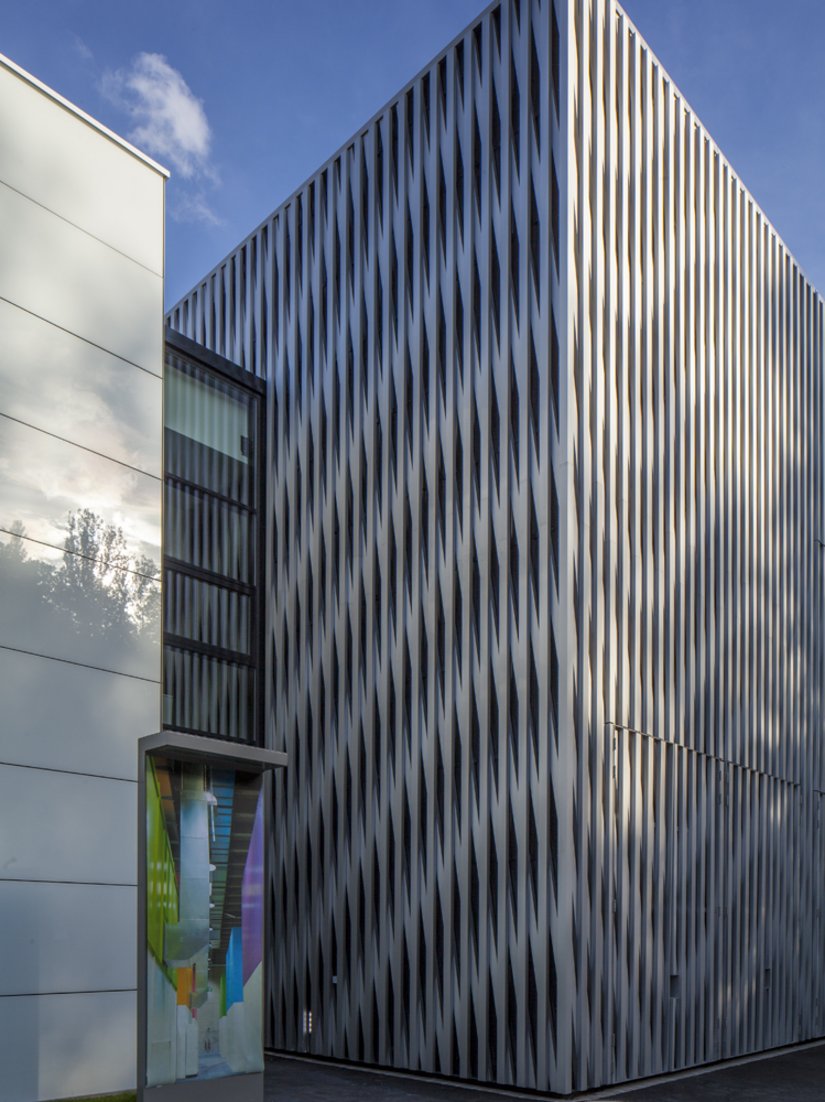 Der L-förmige Bürobau mit horizontalen Fensterbändern umschließt die Aluminiumfassade der Halle.