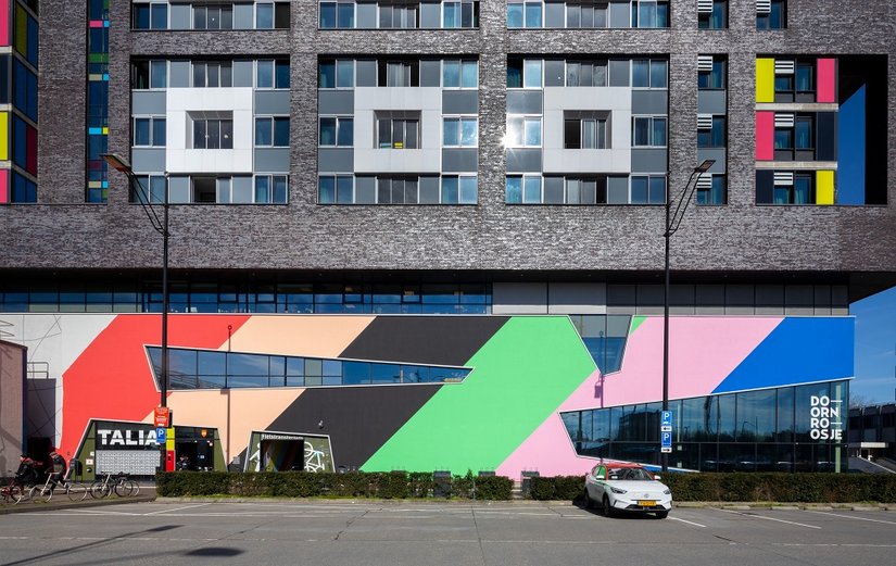 Ein Design mit Botschaft: Zehn leuchtende Farben in diagonaler Ausrichtung prägen die Sockelgeschosse nun von allen Blickrichtungen aus. Das einst graue Kellergeschoss wirkt jetzt wie eine grafische Bühne, die Toleranz, Offenheit und kreative Energie sichtbar macht.